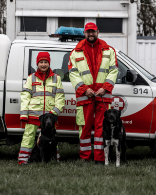 zwei Rettungshundeführer:innen in Uniformen mit roter Haube bzw. Kapperl, zwei Hunde an Leine, stehen vor Samariterbund Auto mit Logo