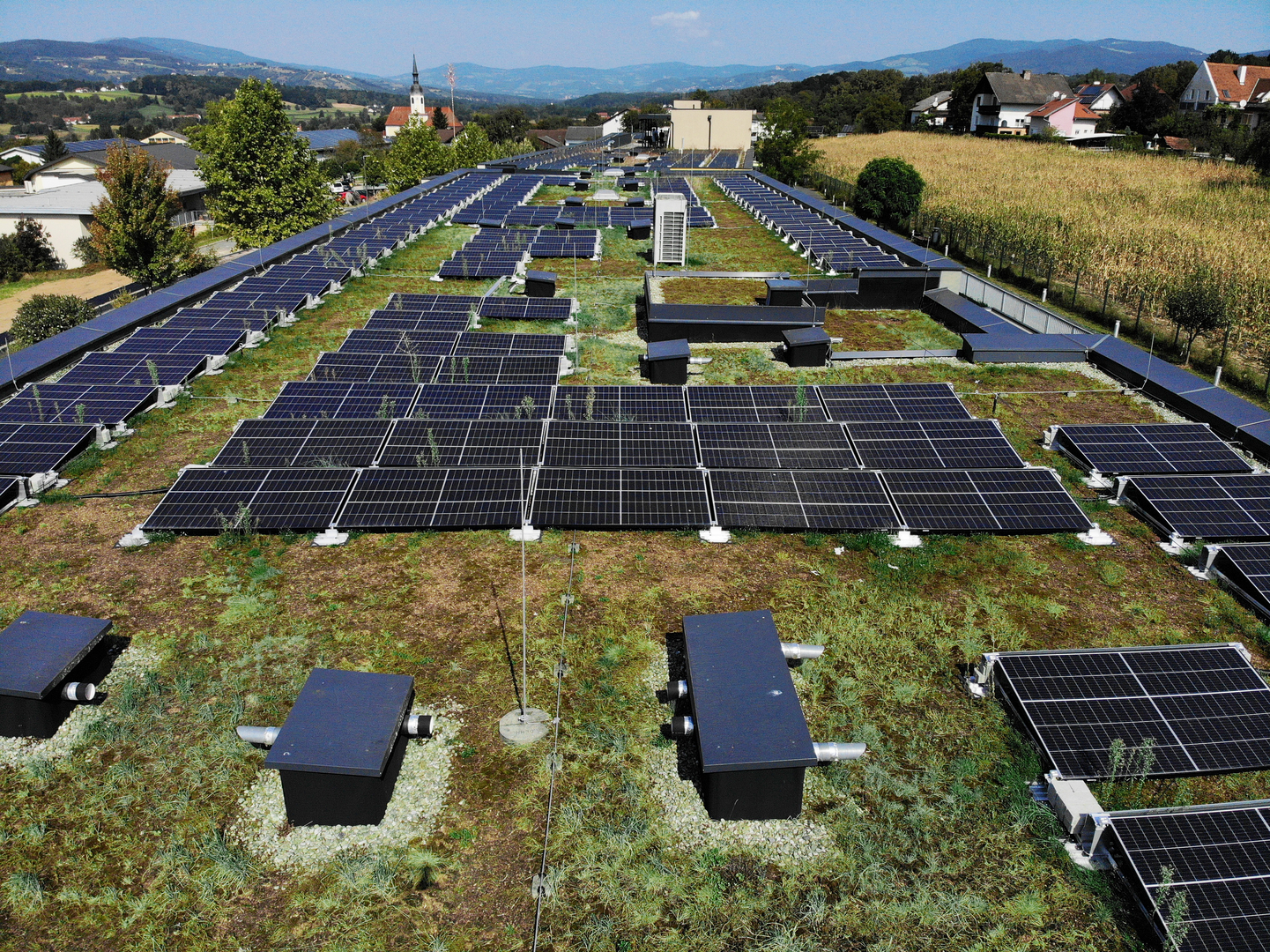 Die Photovoltaikanlage am Dach des Pflegekompetenzzentrums von oben aufgenommen. Sie besteht aus mehreren Solarmodulen. Die Module sind in Reihen angeordnet, umgeben von Gras und anderen Elementen.