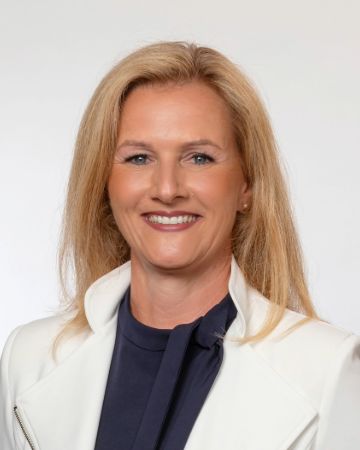 Porträt einer lächelnden Frau mit blonden Haaren, die einen weißen Blazer und ein dunkles Shirt trägt. Der Hintergrund ist neutral.