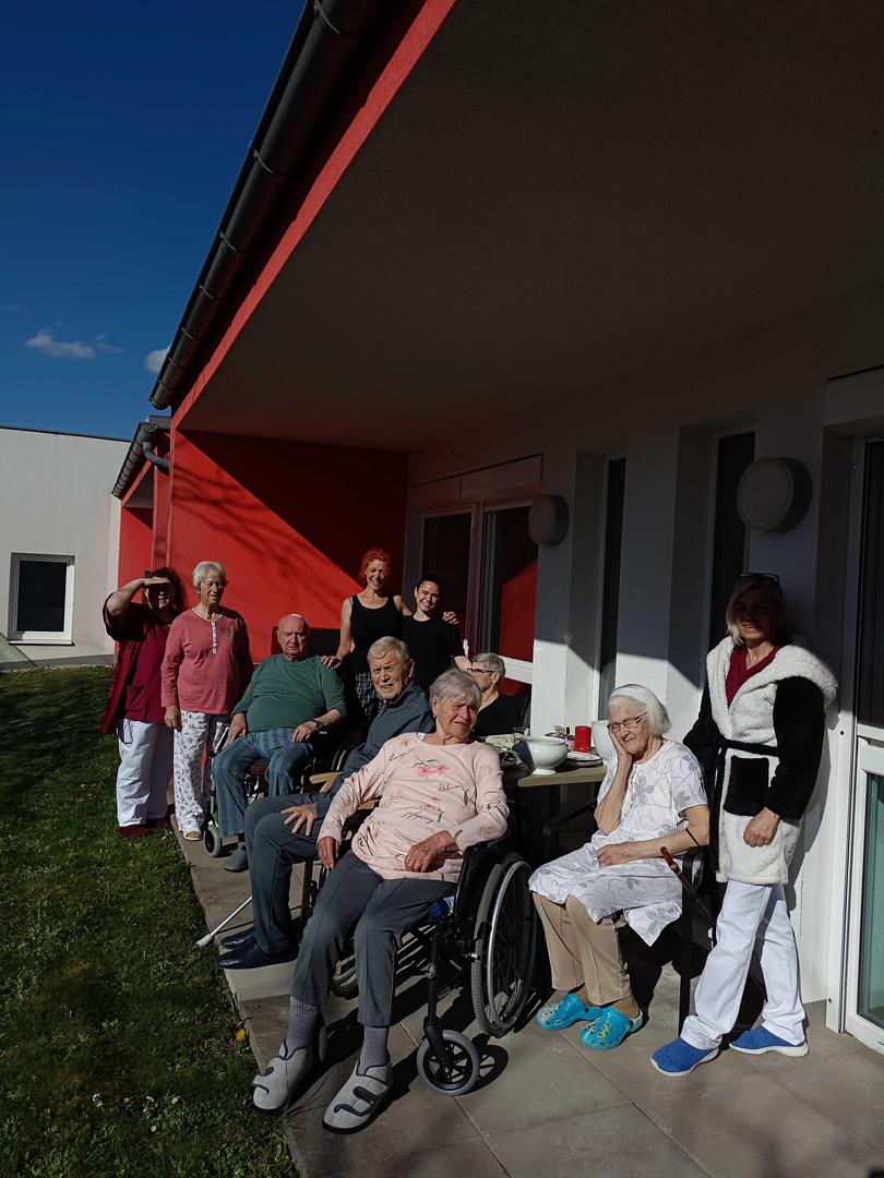 Eine Gruppe von acht älteren Menschen und einer Pflegerin steht vor einem roten Gebäude. Sie lächeln und posieren für das Foto. Einige sitzen im Rollstuhl, während andere stehen. Der Himmel ist blau und die Szene wirkt freundlich und einladend.