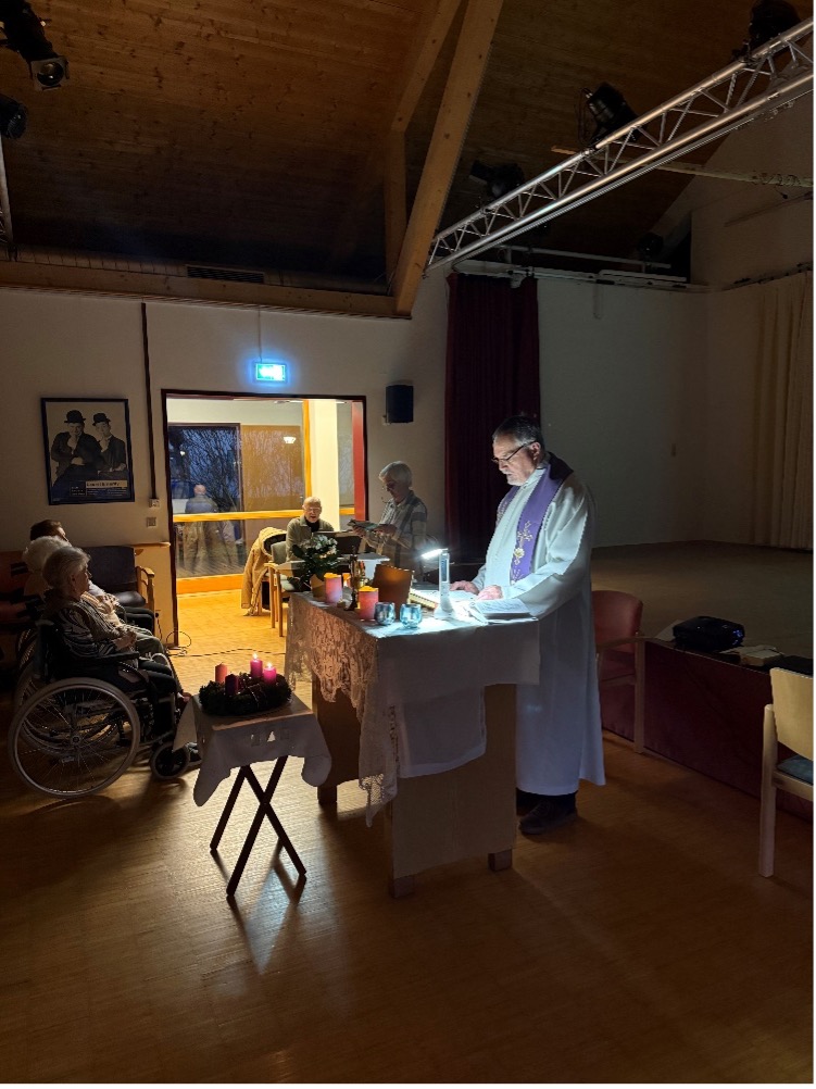 Ein Priester leitet eine Feier mit Kerzenlicht und Teilnehmern in einem Raum.