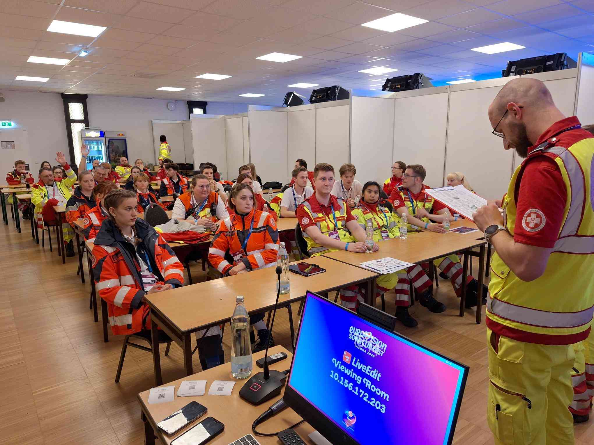 Eine Gruppe von Rettungssanitätern und Helfern sitzt in einem Raum mit Tischen. Sie tragen orangefarbene und gelbe Uniformen. Ein Redner steht vorne und spricht zu ihnen. Auf einem Monitor im Vordergrund sind Informationen zu sehen.