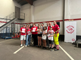Eine Gruppe von Menschen in roten T-Shirts steht in einem Raum. Sie halten Urkunden und Medaillen in den Händen. Im Hintergrund sieht man eine Treppe und ein Banner mit dem Schriftzug „Samma engagiert“. Es sind Kinder und Erwachsene zu sehen, die lächeln.