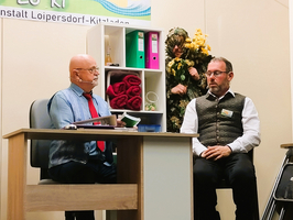 Zwei Männer sitzen an einem Schreibtisch in einem Raum. Einer mit einer Glatze und Brille spricht, der andere trägt eine Weste und sieht konzentriert aus. Im Hintergrund ist eine Person in camouflageähnlicher Kleidung versteckt, die mit Pflanzen dekoriert ist.