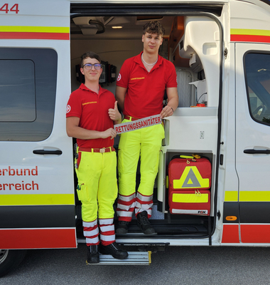 Zwei Rettungssanitäter stehen an einem Rettungswagen und zeigen Daumen hoch.