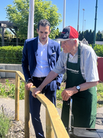 Ein junger Mann in einem Anzug steht neben einem älteren Herren, der eine Schürze trägt und mit einem Rollator steht. Sie schauen sich gemeinsam einen Gartenbereich an, umgeben von Pflanzen und einem Zaun.