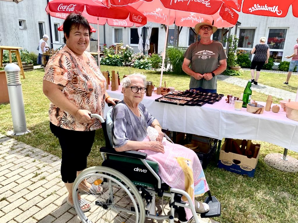 Eine ältere Frau im Rollstuhl steht neben einer lächelnden Frau an einem Marktstand.