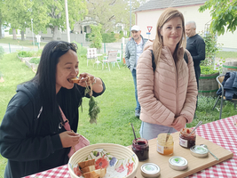 Zwei Frauen stehen an einem gedeckten Tisch im Freien. Eine Frau probiert gerade ein Essen aus einer Schüssel, während die andere lächelt und beobachtet. Im Hintergrund sind weitere Personen und eine grüne Landschaft zu sehen.