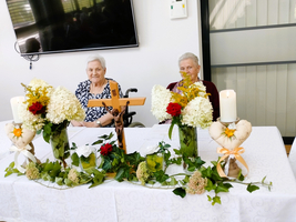 Zwei ältere Frauen sitzen an einem Tisch mit Blumen und einem Kreuz.