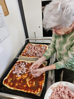 Eine ältere Frau belegt frische Pizza mit gewürfeltem Schinken.