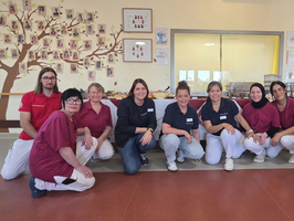 Eine Gruppe von acht Personen in Arbeitskleidung posiert gemeinsam in einem Raum. Sie sitzen und stehen auf einer gepflasterten Fläche, umgeben von einer Wand mit Fotografien. Im Hintergrund sind Tische mit Essen sichtbar. Die Atmosphäre wirkt freundlich und einladend.