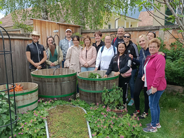 Eine Gruppe von etwa 15 Personen steht in einem Garten, umgeben von Holzbalken und Pflanzen. Im Vordergrund befindet sich ein Weg aus Gras und Erde. Die Teilnehmer lächeln und posieren gemeinsam, während Bäume und einige Häuser im Hintergrund sichtbar sind.