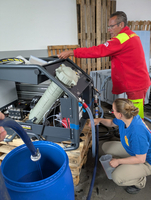 Zwei Personen arbeiten an einer Maschine, während eine Flüssigkeit in einen Eimer fließt.