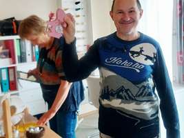 Ein lächelnder Mann hält ein rosa Bastelobjekt in der Hand, während im Hintergrund eine Frau beschäftigt ist. Im Raum sind Bastelmaterialien und eine bunte Dekoration mit dem Wort „NAH“.