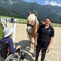Eine ältere Frau im Rollstuhl spricht mit einem Mädchen und einem Pferd.