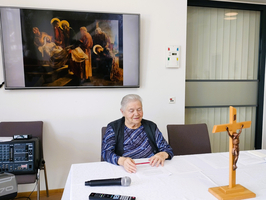 Eine ältere Frau sitzt an einem Tisch mit einem Mikrofon und Blick auf eine Leinwand mit einem Gemälde im Hintergrund. Auf dem Tisch steht eine Holzfigur eines gekreuzigten Christus. Der Raum ist hell und modern eingerichtet.