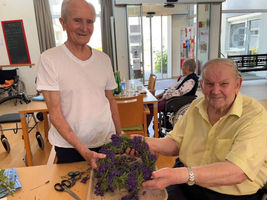 Zwei Männer halten gemeinsam einen Kranz aus Lavendel in einem Aufenthaltsraum.