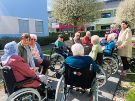 Eine Gruppe von älteren Menschen in Rollstühlen sitzt im Freien. Einige Personen lächeln und unterhalten sich, während sie von Bäumen und Blumen umgeben sind. Eine Person steht hinter der Gruppe und scheint freundlich mit ihnen zu interagieren.