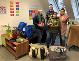 vier Personen mit selbstgestrickten Hauben, stehen nebeneinander in einem Raum, davor Sackerl voller selbstgestrickter Hauben und Schals, im Hintergrund Plakate, Pflanzen