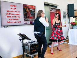 Ein Sängerpaar steht auf einer Bühne. Der Mann trägt eine schwarze Weste und hält ein Mikrofon, während die Frau in einem roten Kleid mit weißen Punkten singt. Im Hintergrund hängt ein Banner mit einer Dankesbotschaft an die Sponsoren. Ein Keyboard ist auf der linken Seite sichtbar.