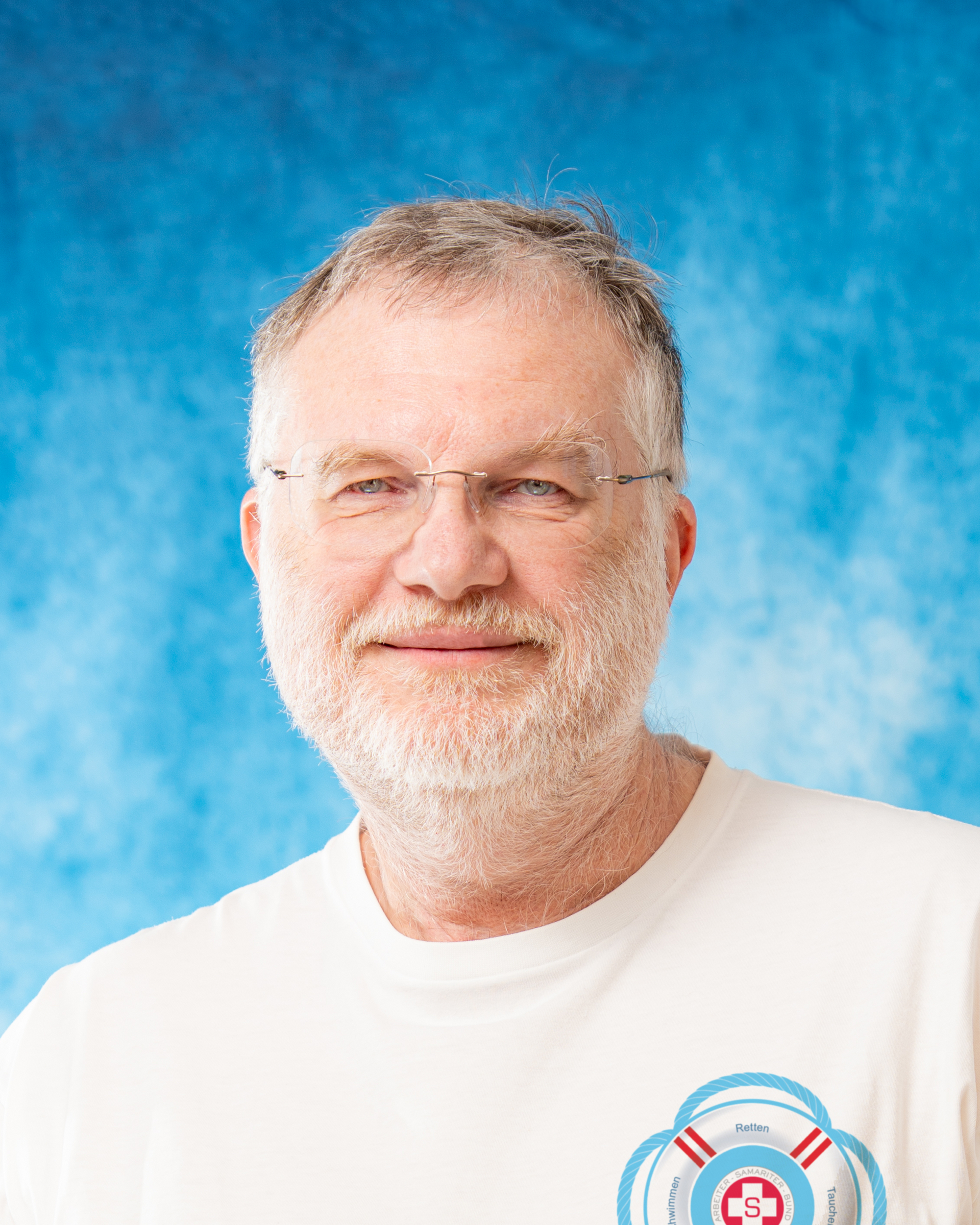 Ein Mann mit Brille und Bart lächelt vor einem blauen Hintergrund.