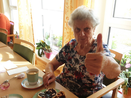 Ältere Frau mit Daumen hoch, vor ihr ein Stück Kuchen und ein Kaffee.