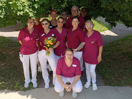 Eine Gruppe von zwölf Frauen in roten Arbeitsoberteilen und weißen Hosen steht unter großen, grünen Bäumen. Einige Frauen halten Blumen in der Hand. Sie lächeln und posieren für das Foto, das in einer sonnigen Umgebung aufgenommen wurde.