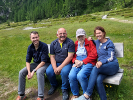 Vier Personen sitzen auf einer Bank in der Natur, umgeben von Bäumen und Wiesen.