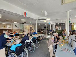 Eine lebhafte Seniorenresidenz mit mehreren Gästen, die an Tischen sitzen und essen. Einige Personen benutzen Rollstühle, während andere aktiv in Gespräche vertieft sind. Der Raum ist hell und freundlich dekoriert, mit Luftballons und Pflanzen im Hintergrund.