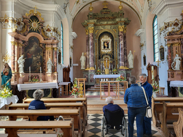 Innenansicht einer Kirche mit zwei Personen, die miteinander sprechen, während eine andere Person im Rollstuhl sitzt. Der Altar im Hintergrund ist reich verziert und umgeben von Statuen und Blumenarrangements. Bänke sind im Vordergrund sichtbar.