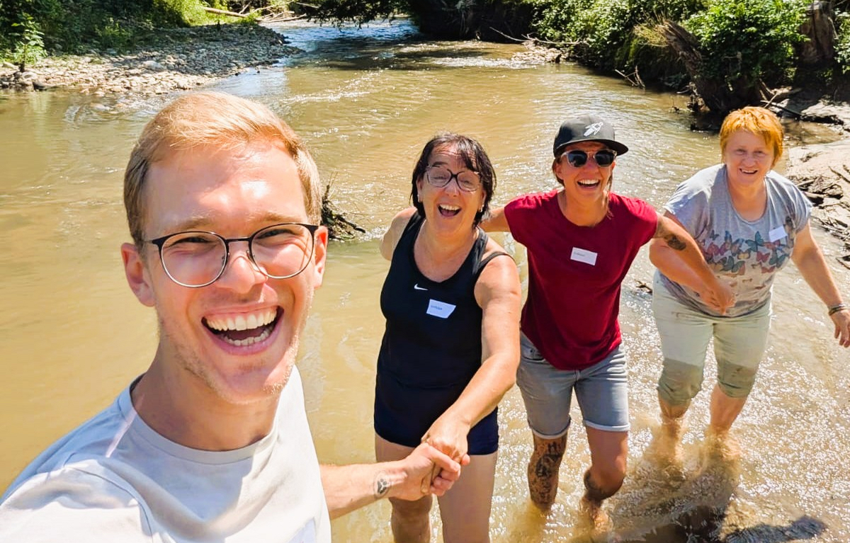 Eine Gruppe von vier Personen steht im flachen Wasser eines Baches. Sie lachen und halten sich in einer Kette an den Händen. Im Hintergrund sind Bäume und Ufervegetation sichtbar. Es ist ein sonniger Tag.