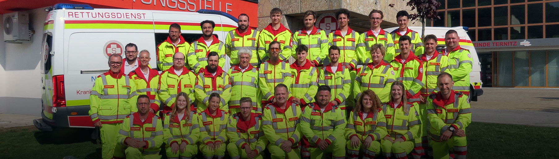 Gruppenfoto von Rettungsdienstmitarbeitern in auffälligen Uniformen vor einem Rettungswagen.