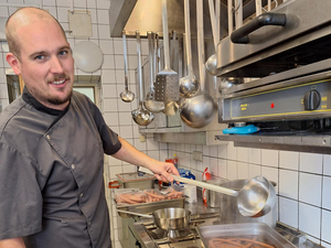 Koch Tobias vom Samariter Suppentopf in Küche hält Schöpflöffel und blickt in Kamera, daneben ein großes Behältnis mit Frankfurter Würsten in Wasser, im Hintergrund Küchenausstattung