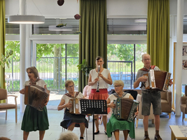 Eine Gruppe von fünf Musikern spielt in einem Raum. Zwei Frauen spielen Akkordeon, eine Frau spielt Klarinette, während zwei weitere Musiker mit Akkordeons stehen. Sie tragen farbenfrohe Kleidung und sind umgeben von einem informellen, einladenden Ambiente.