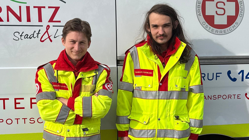 Zwei Personen in auffälligen Sicherheitsjacken stehen vor einem Fahrzeug mit dem Logo eines Rettungsdienstes. Beide tragen rot-gelbe Kleidung, die für Notfall- und Rettungsdienste typisch ist. Der Hintergrund zeigt die Seite des Fahrzeugs.