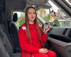 Eine Frau im roten Outfit hält ein Funkgerät in einem Fahrzeug.