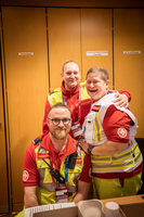 Drei Rettungskräfte in Uniform stehen und lachen vor einem Holzregal. Zwei Personen stehen im Hintergrund, eine Person kniet vorne. Alle tragen rote und gelbe Kleidung mit Einsatzabzeichen.