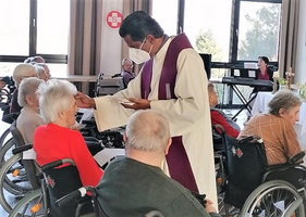 Ein Priester segnet eine ältere Frau in einem Raum voller Rollstühle.