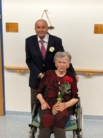 Ein älteres Paar, der Mann im Anzug, die Frau im Rollstuhl mit einer Blume.