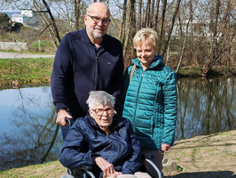 Drei Personen stehen am Ufer eines Gewässers. Eine ältere Frau sitzt im Rollstuhl, während zwei Personen, ein Mann und eine Frau, hinter ihr stehen. Im Hintergrund sind Bäume und ein Schild sichtbar. Es ist sonnig und die Umgebung wirkt freundlich.