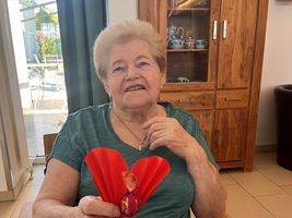 Eine ältere Frau sitzt an einem Tisch und hält eine rote Blume oder eine Dekoration in der Hand. Sie trägt ein grünes Oberteil und lächelt, während sie in die Kamera schaut. Im Hintergrund ist ein Holzschrank und Fenster mit Tageslicht sichtbar.