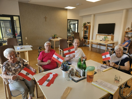 Eine Gruppe von vier Senioren sitzt an einem Tisch in einem Gemeinschaftsraum. Jeder hält eine kleine Flagge. Im Hintergrund sind weitere Personen und Möbel zu sehen. Der Raum hat eine helle, freundliche Atmosphäre.