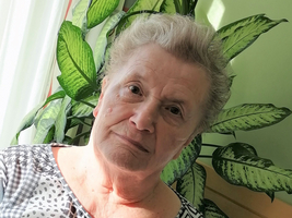Ältere Frau mit nachdenklichem Ausdruck, im Hintergrund temperamentvolle grüne Pflanzen.