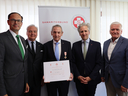 Fünf Männer stehen in einem Raum und posieren für ein Foto. Der Mann in der Mitte trägt eine Auszeichnung und hält ein Zertifikat in der Hand. Im Hintergrund ist ein Banner des Samariterbundes zu sehen.