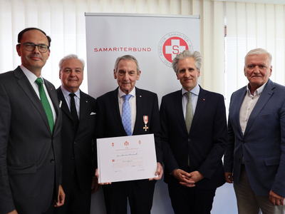 Fünf Männer stehen in einem Raum und posieren für ein Foto. Der Mann in der Mitte trägt eine Auszeichnung und hält ein Zertifikat in der Hand. Im Hintergrund ist ein Banner des Samariterbundes zu sehen.