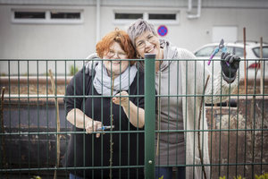 Zwei Frauen lächeln hinter einem Zaun und halten Gartenschneider.
