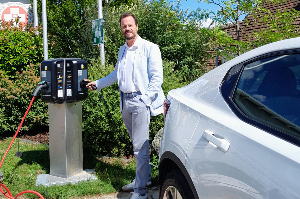 Andreas Balog steht neben einer Ladestation für Elektrofahrzeuge. Im Hintergrund sind grüne Pflanzen und die Samariterbund-Fahne zu sehen. Ein weißes Elektroauto steht im Vordergrund und wird gerade aufgeladen.
