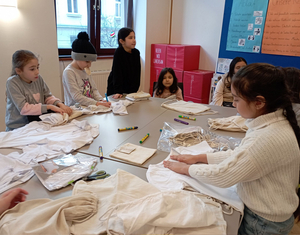Eine Gruppe Mädchen sitzt rund um einen Tisch, vor ihnen weiße Tshirts, die künstlerisch gestaltet werden sollen.