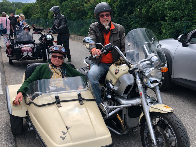 Ein Mann sitzt auf einem Motorrad mit Beiwagen, in dem eine Frau mit Sonnenbrille und einem Hut sitzt. Beide lächeln. Im Hintergrund sind weitere Motorräder zu sehen und eine grüne, bewaldete Umgebung.