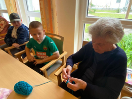 Eine ältere Frau strickt, während zwei Jungen ihr aufmerksam zuschauen.